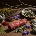 Bâton de fumigation sauge sang de dragon, feuilles rouge vif ficelées de blanc, posé sur table en bois avec herbes et fleurs violettes.