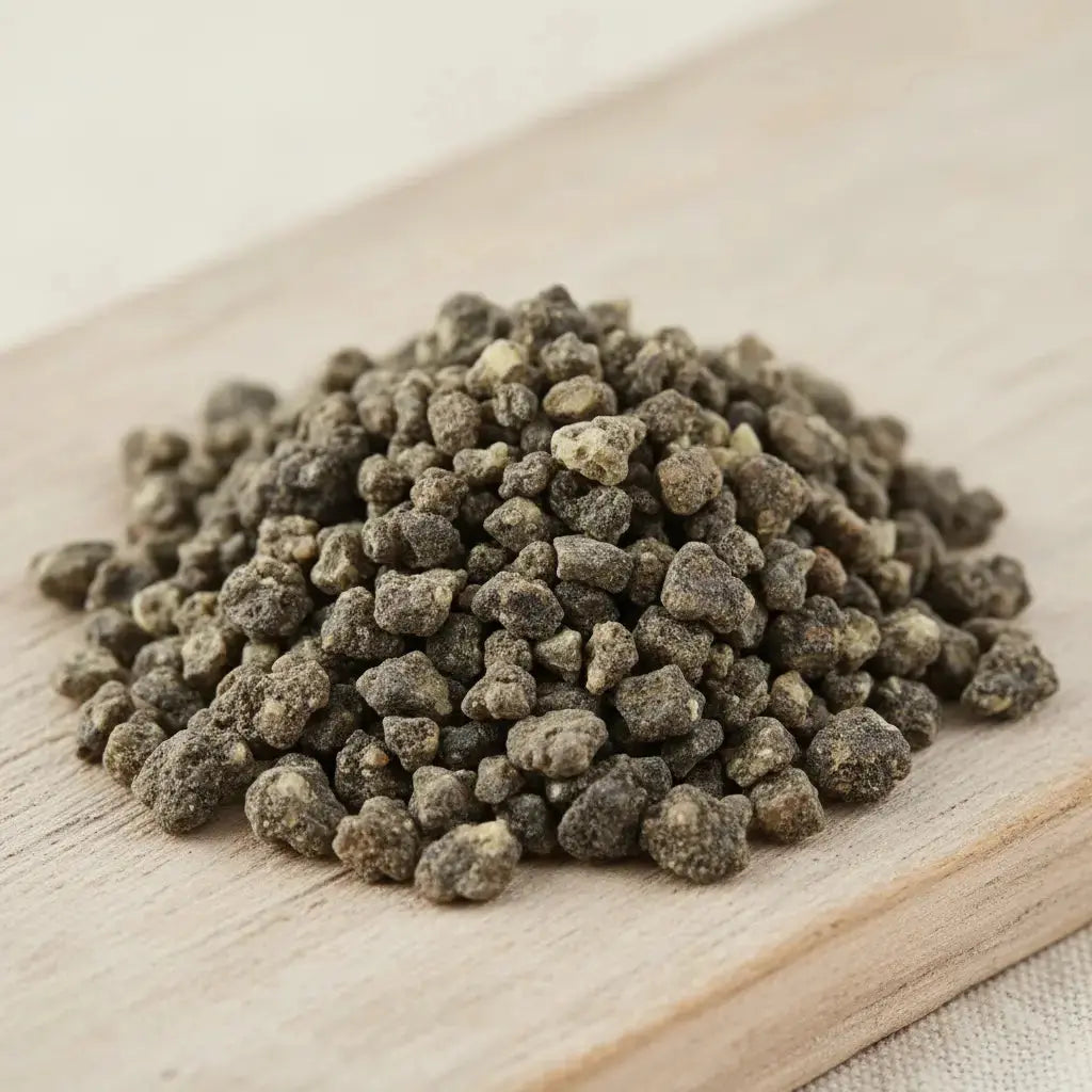 Gros plan sur un monticule de grains de résine d'Encens Benjoin de Sumatra, montrant des morceaux irréguliers de couleur gris-brun foncé/terreux, sur une planche de bois clair.