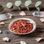 Bois de santal rouge pour fumigation, copeaux rouge-orangé dans une coquille d’ormeau sur table en bois avec pétales et cristaux.