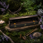 Boîte Porte Encens en Bois Noir - Om Doré entrouverte sur un socle en bois moussue, dévoilant ses coupelles dorées intérieures à côté de cônes d'encens.