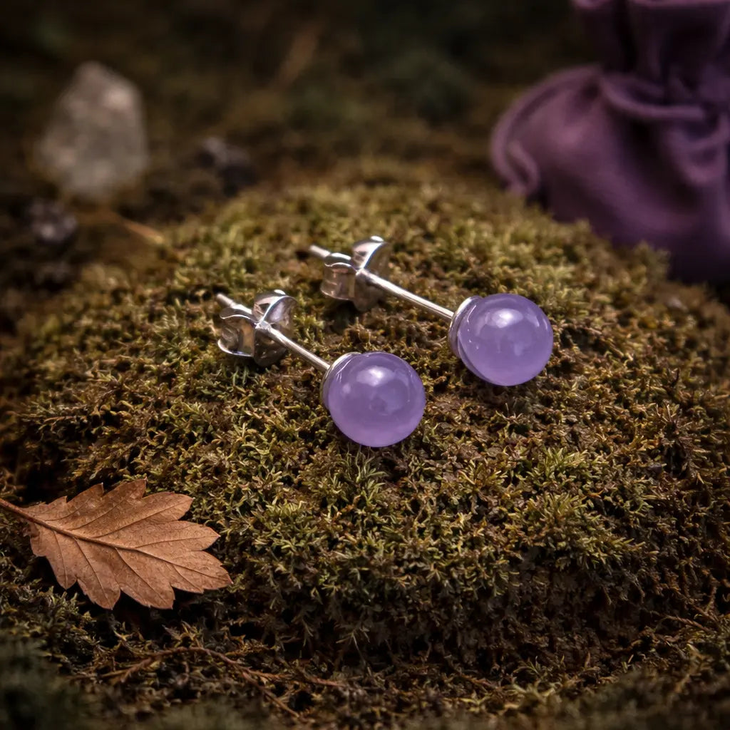 Paire de clous d'oreilles ornés de perles d'améthyste violettes et translucides, posées sur une mousse épaisse à côté d'une feuille de chêne séchée.
