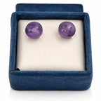 Une paire de boucles d'oreilles puces en améthyste violette, délicatement posées sur un amas de cristal de roche brut blanc, entouré de grandes feuilles vertes.