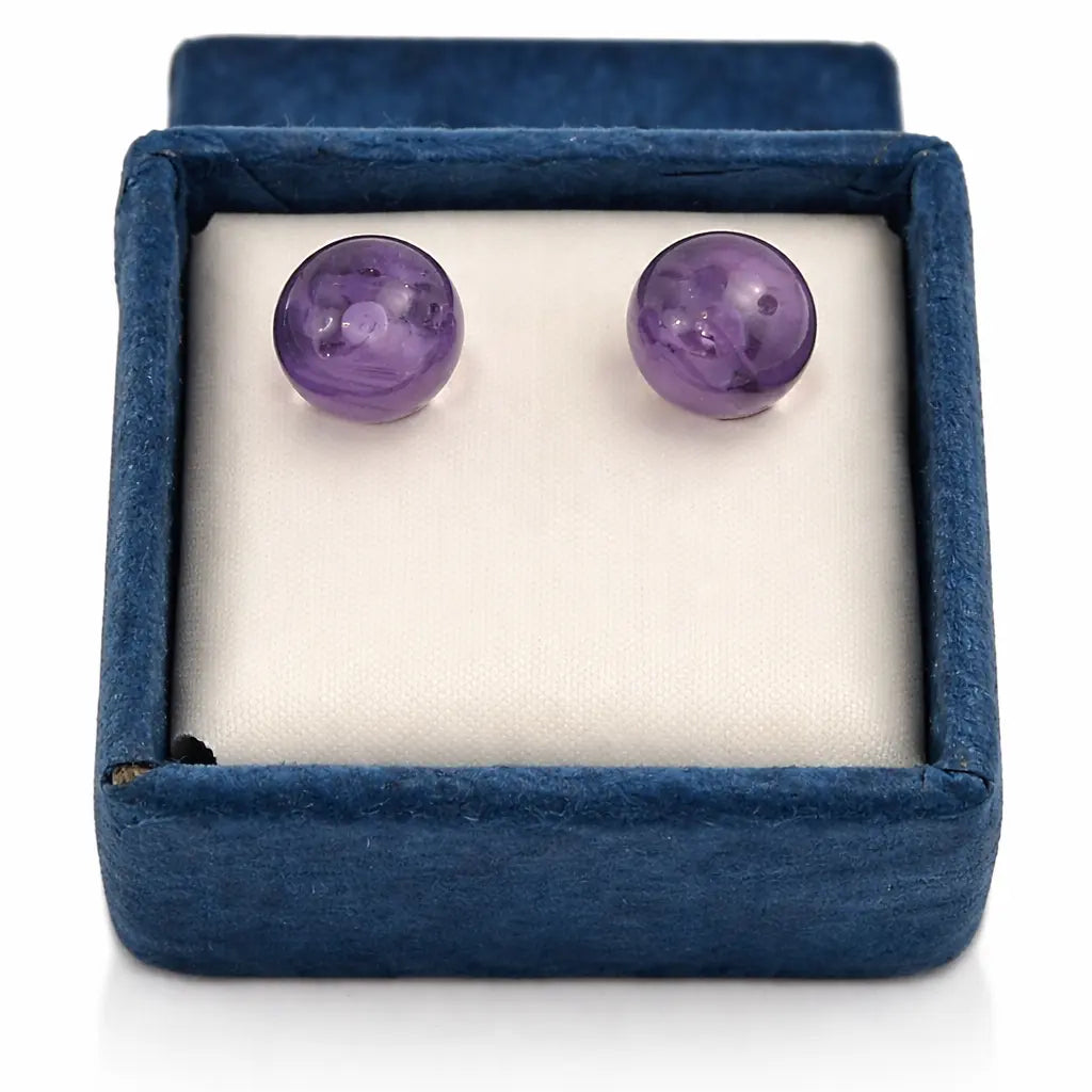 Une paire de boucles d'oreilles puces en améthyste violette, délicatement posées sur un amas de cristal de roche brut blanc, entouré de grandes feuilles vertes.