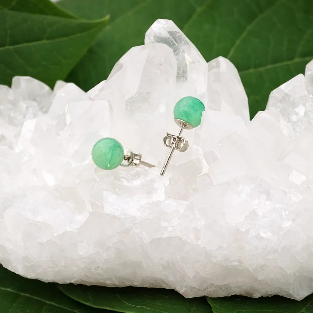 Une paire de boucles d'oreilles puces en aventurine verte, délicatement posées sur un amas de cristal de roche brut blanc, avec des feuilles vertes en arrière-plan.