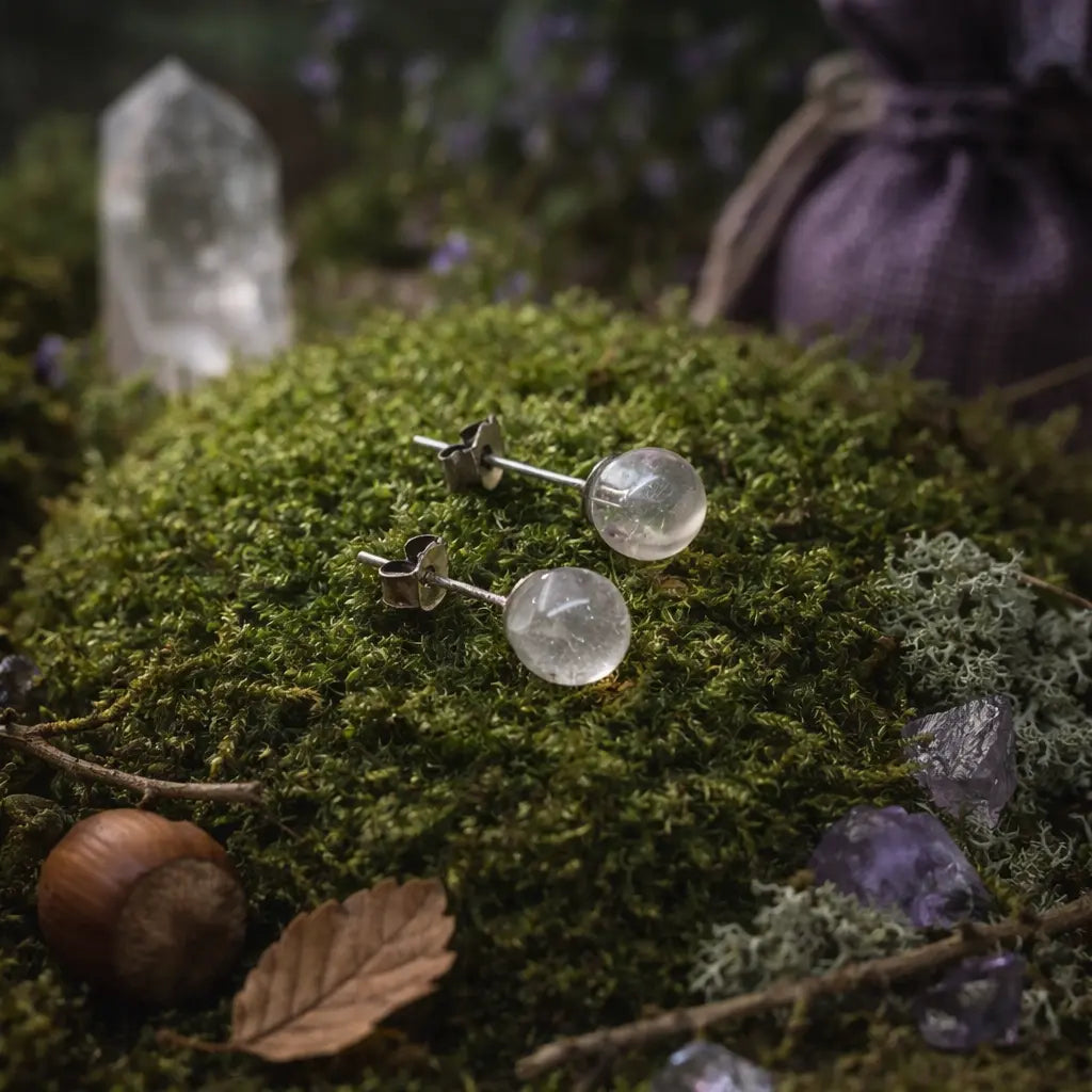 Clous d'oreilles ornés de perles de cristal de roche pur et translucide, déposés sur un tapis de mousse verte parsemé de petits cristaux bruts.