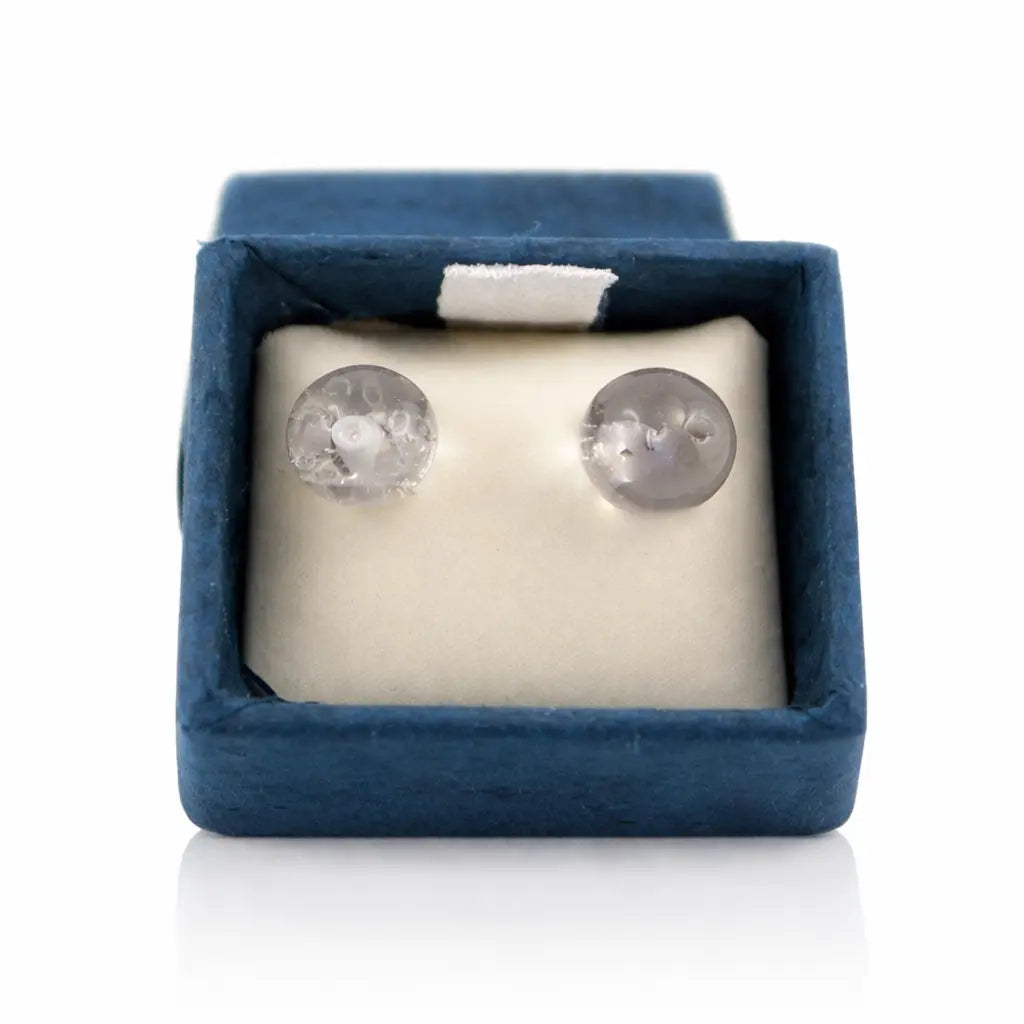 Une paire de boucles d'oreilles puces en cristal de roche, présentant deux perles transparentes aux inclusions naturelles, soigneusement disposées dans leur écrin bleu sur un coussinet blanc.