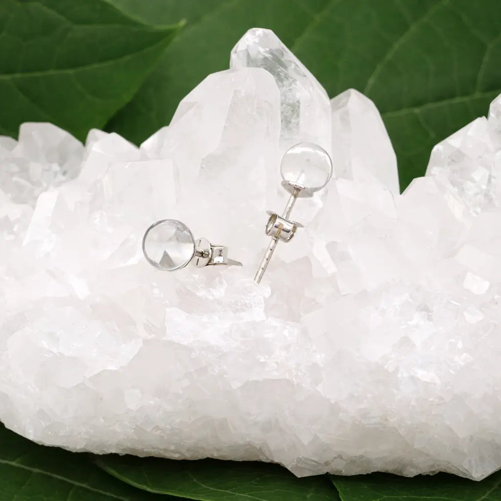 Une paire de boucles d'oreilles puces en cristal de roche, délicatement posées sur un amas de cristal de roche brut blanc, le tout agrémenté de feuilles vertes en arrière-plan.