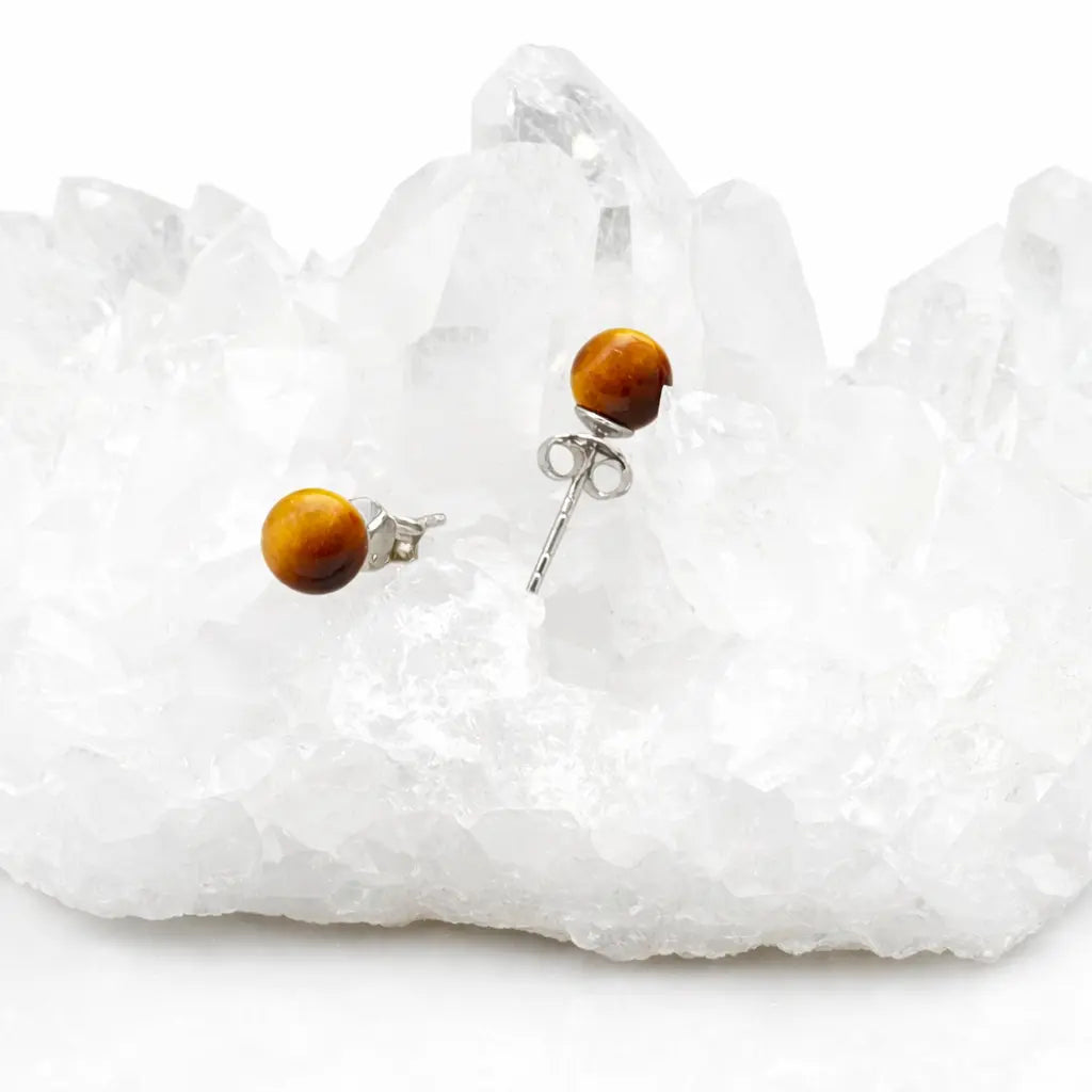 Une paire de boucles d'oreilles puces en œil de tigre, aux tons ambrés et dorés, délicatement posées sur un amas de cristal de roche brut d'un blanc pur.