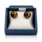 Une paire de boucles d'oreilles puces en œil de tigre, présentant deux perles rondes aux reflets dorés et marron, soigneusement disposées dans leur écrin bleu sur un coussinet blanc.