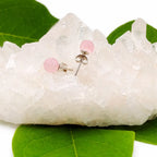 Une paire de boucles d'oreilles puces en quartz rose, délicatement posées sur un amas de cristal de roche brut décoré de quelques feuilles vertes.