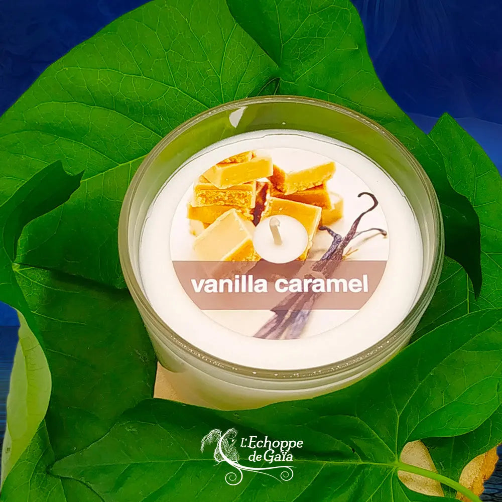 Bougie naturelle parfumée vanille et caramel en verre, avec étiquette illustrée de morceaux de caramel et texte vanilla caramel, posée sur de grandes feuilles vertes.