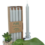 Bougies chandelles “PURE – Natural Wax” : lot de 4 bougies gris clair dans un étui carton, avec une chandelle gris clair présentée à côté dans un bougeoir en verre (mise en scène avec feuilles vertes + petite succulente).