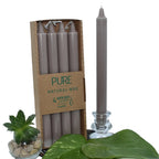 Bougies chandelles “PURE – Natural Wax” : lot de 4 bougies couleur taupe / gris chaud dans un étui carton, avec une chandelle assortie présentée à côté dans un bougeoir en verre (mise en scène avec feuilles vertes + petite succulente).