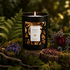 Bougie parfumée Eskalia Hiver à l'étiquette dorée illustrée de feuilles, allumée sur un tronc moussue au milieu de fougères et de fleurs violettes.
