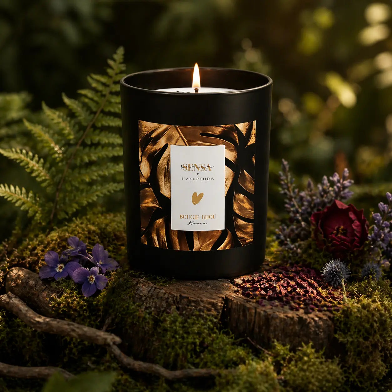 Bougie parfumée Eskalia Hiver à l'étiquette dorée illustrée de feuilles, allumée sur un tronc moussue au milieu de fougères et de fleurs violettes.