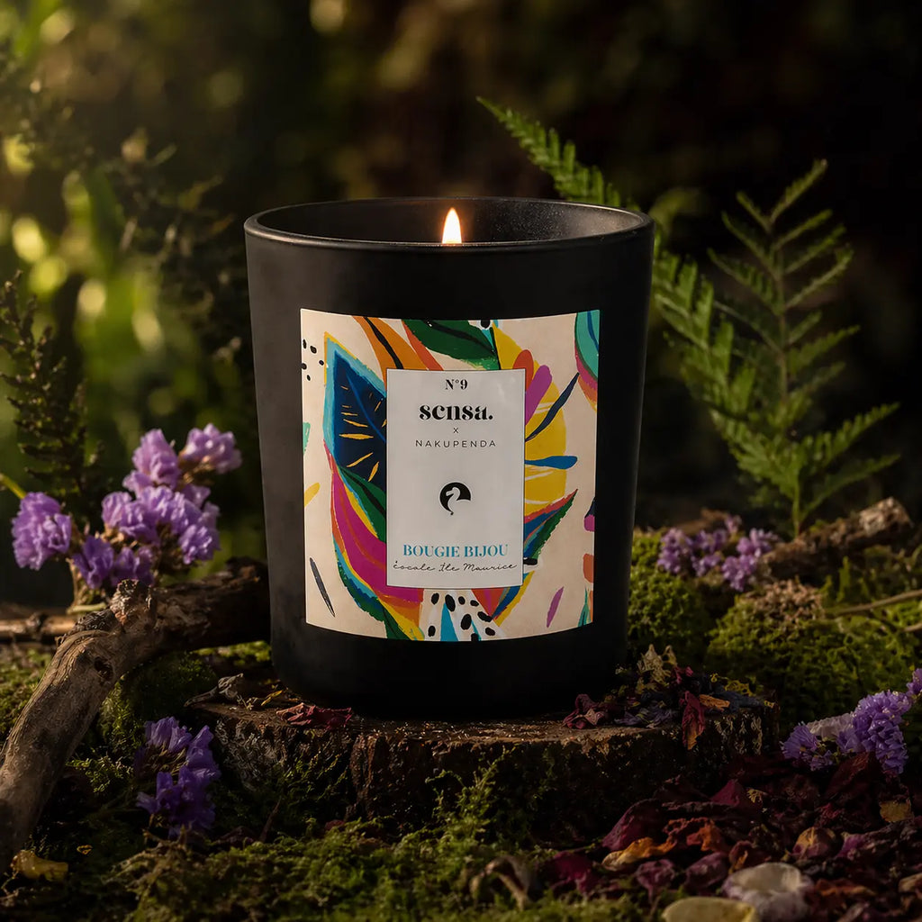 Bougie parfumée Eskalia Escale Ile Maurice allumée sur un socle de bois moussue, entourée de fleurs violettes et de fougères dans une ambiance de sous-bois naturel.