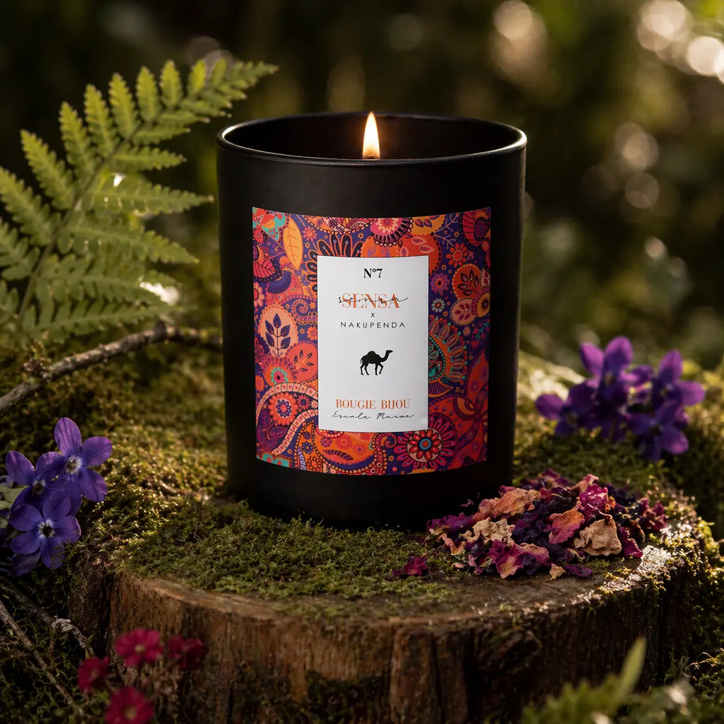 Bougie parfumée Eskalia Escale Maroc à l'étiquette colorée aux motifs orientaux, allumée sur une souche de bois moussue entourée de fleurs violettes et d'une fougère.
