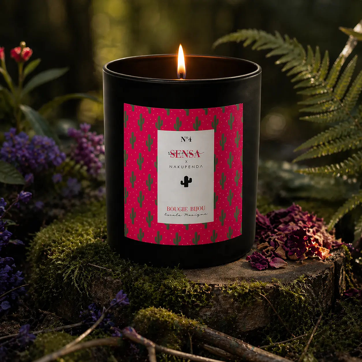 Bougie parfumée Eskalia Escale Mexique à l'étiquette rouge et noire, allumée sur une souche de bois moussue entourée de fleurs violettes et de fougères dans un sous-bois.