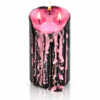 Grande bougie pilier vampire Draculina noire à 3 mèches allumées, laissant couler de la cire rose le long de ses parois, sur fond blanc avec reflet.