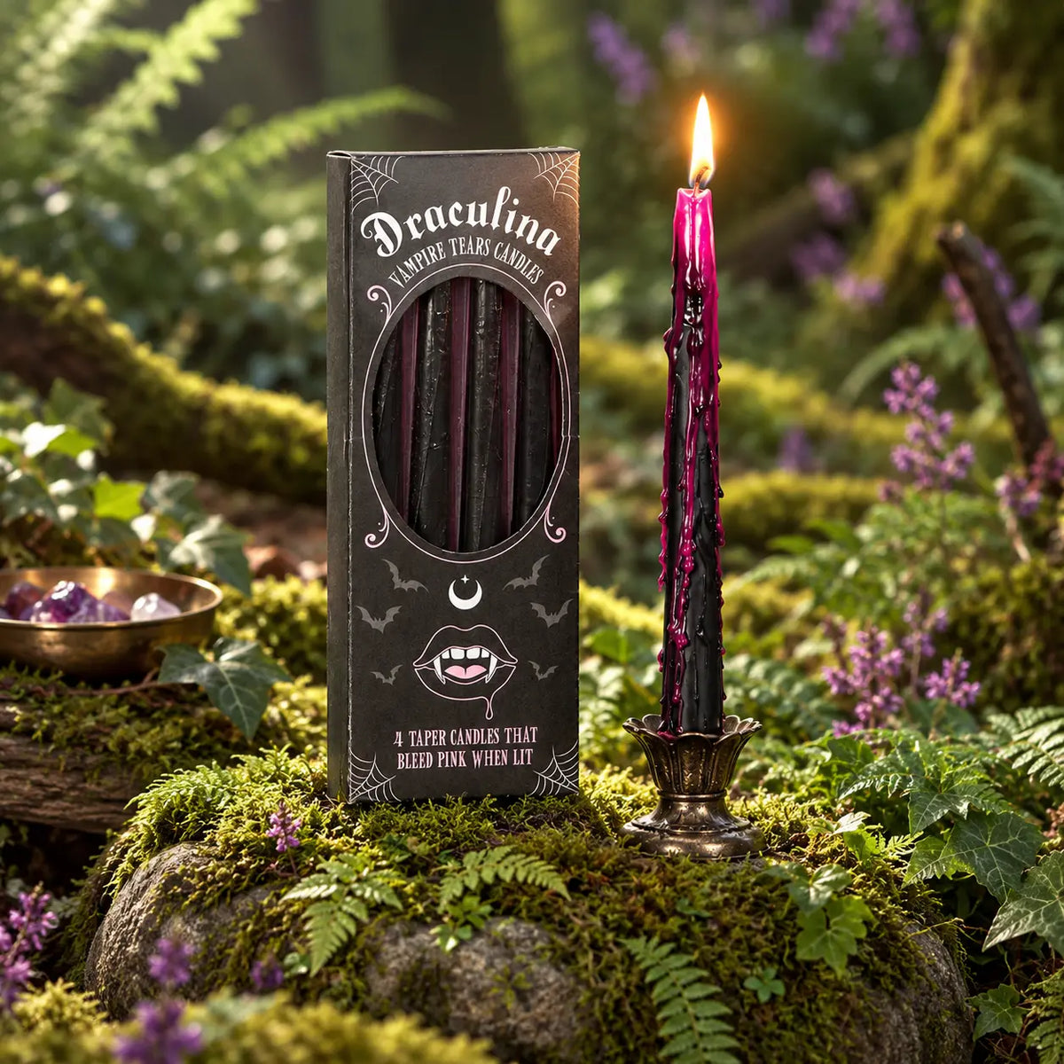 Boîte noire de bougies Draculina Vampire tears candles avec fenêtre montrant 4 cierges effilés, posée sur de la mousse à côté d'un cierge allumé aux coulures roses sur bougeoir en métal.