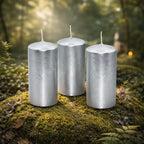 Trio de bougies piliers métalisées cylindre argent de 10 cm de haut disposées sur de la mousse en forêt.