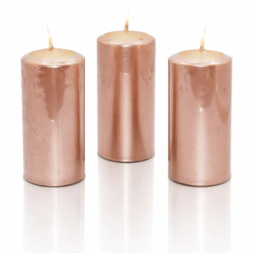 Un lot de trois bougies piliers métallisées de couleur or rose (ou vieux rose), de forme cylindrique, présentées debout sur un fond blanc avec un reflet à leur base.
