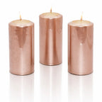 Un lot de trois bougies piliers métallisées de couleur or rose (ou vieux rose), de forme cylindrique, présentées debout sur un fond blanc avec un reflet à leur base.