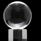 Boule de cristal très transparente, photographiée sur fond noir : la sphère capte de forts reflets lumineux et repose sur un socle cubique en verre/plexi clair.