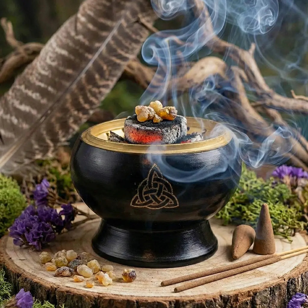 Brûle-encens en métal noir et doré avec motif celte gravé, charbon incandescent fumant et grains de résine, posé sur rondin en extérieur.