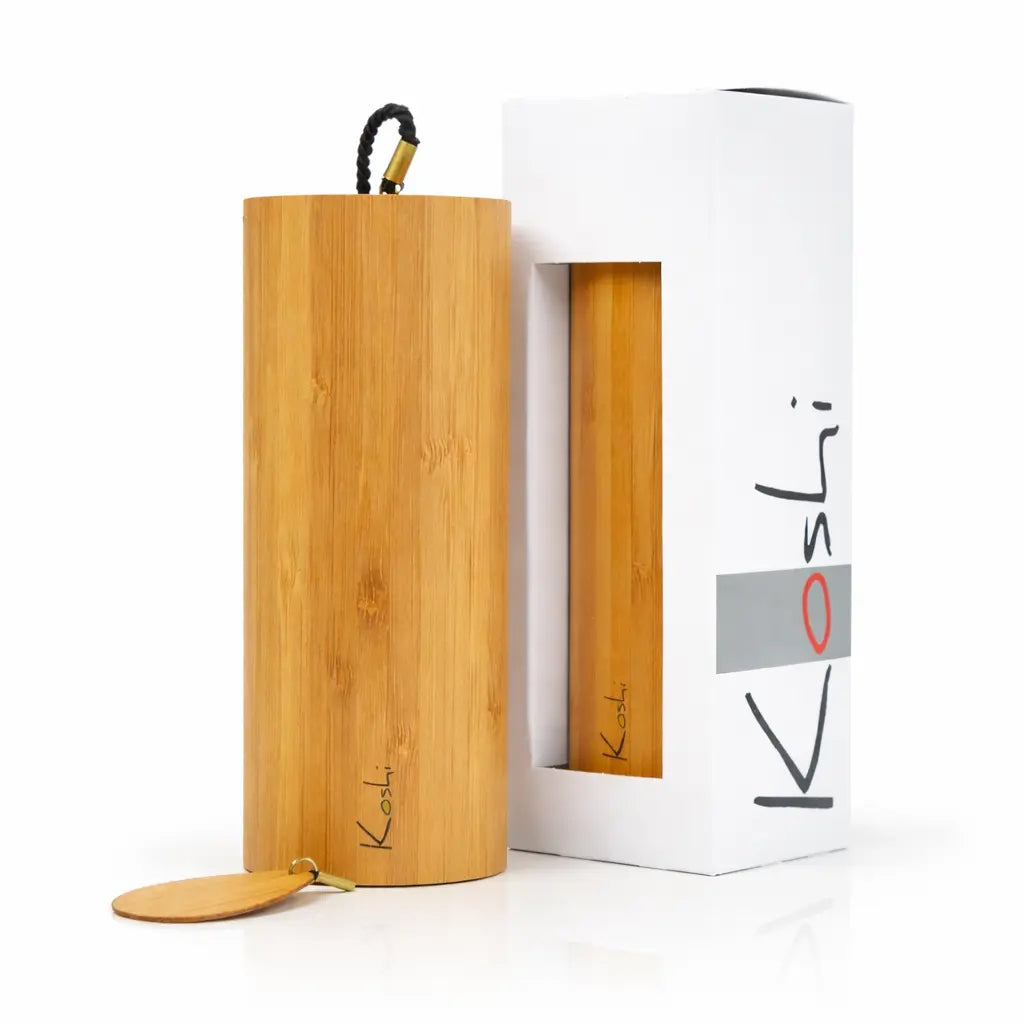 Un carillon Koshi Ignis, reconnaissable à son point rouge sur le logo, présenté debout à côté de son emballage d'origine. Le tube en bambou naturel est accompagné de son battant en bois et de sa corde d'attache noire, le tout sur un fond blanc réfléchissant.