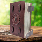Un carnet en cuir artisanal marron doté de deux lanières de fermeture, orné en son centre d'une pierre d'oeil de tigre sertie dans une tresse de cuir, présenté sur un support en bois.