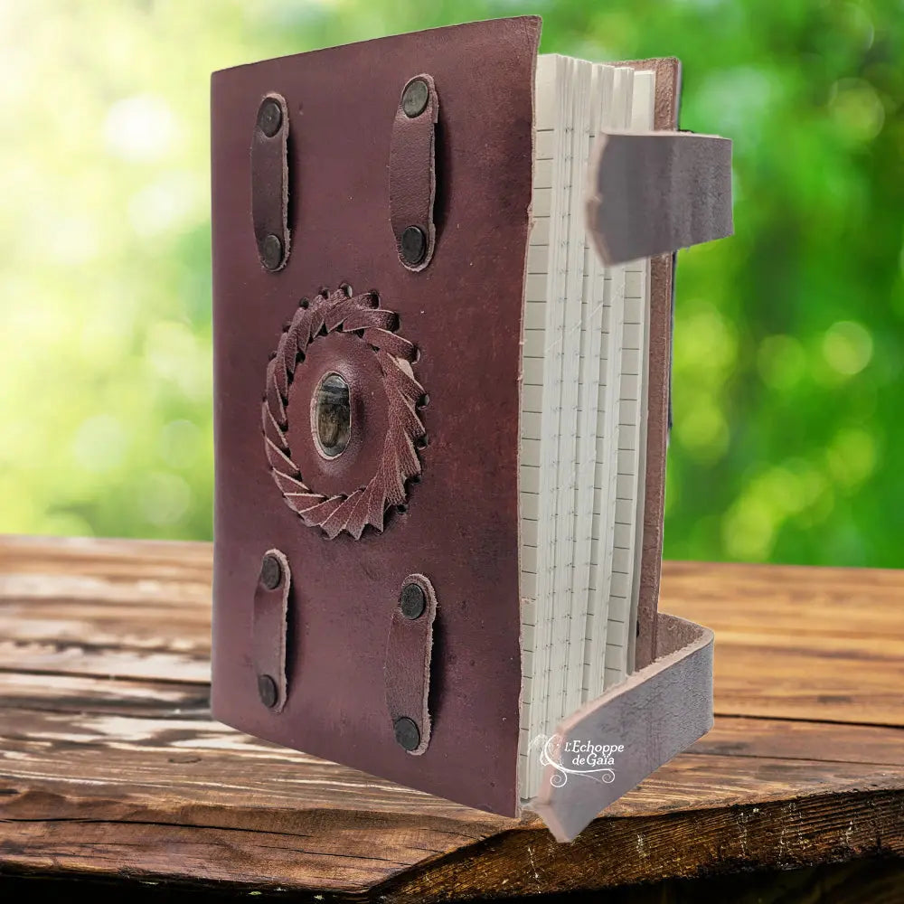 Un carnet en cuir artisanal marron doté de deux lanières de fermeture, orné en son centre d'une pierre d'oeil de tigre sertie dans une tresse de cuir, présenté sur un support en bois.
