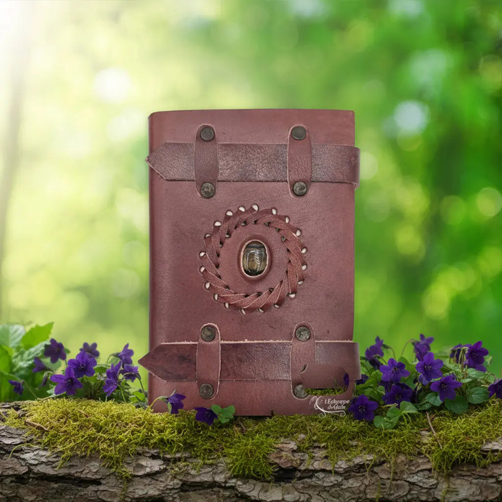 Carnet Ceinturé en Cuir Artisanal - Oeil de Tigre fermé par deux lanières, orné d'une pierre centrale, posé sur une branche moussue avec des fleurs violettes dans une forêt.