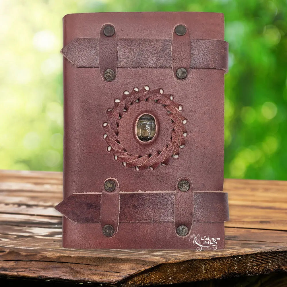 La couverture d'un carnet en cuir artisanal marron fermé par deux lanières horizontales, ornée d'une pierre d'œil de tigre centrale sertie dans une tresse de cuir, sur un support en bois.