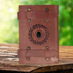 La couverture d'un carnet en cuir artisanal marron fermé par deux lanières horizontales, ornée d'une pierre d'onyx noire centrale sertie dans une tresse de cuir, sur un support en bois.
