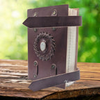 Un carnet en cuir artisanal marron doté de deux lanières de fermeture, orné en son centre d'une pierre de lune sertie dans une tresse de cuir, présenté sur un support en bois.