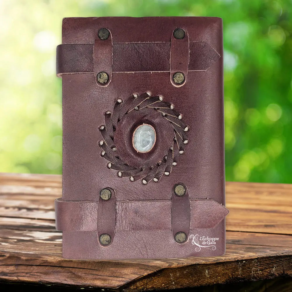 La couverture d'un carnet en cuir artisanal marron fermé par deux lanières horizontales, ornée d'une pierre de lune centrale sertie dans une tresse de cuir, sur un support en bois.