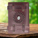 La couverture d'un carnet en cuir artisanal marron fermé par deux lanières horizontales, ornée d'une pierre de lune centrale sertie dans une tresse de cuir, sur un support en bois.