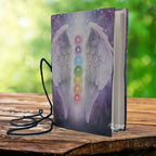 Un carnet de notes à couverture souple illustrée d'ailes d'ange et des symboles des sept chakras sur un fond violet étoilé, accompagné d'un cordon de fermeture noir sur un support en bois.