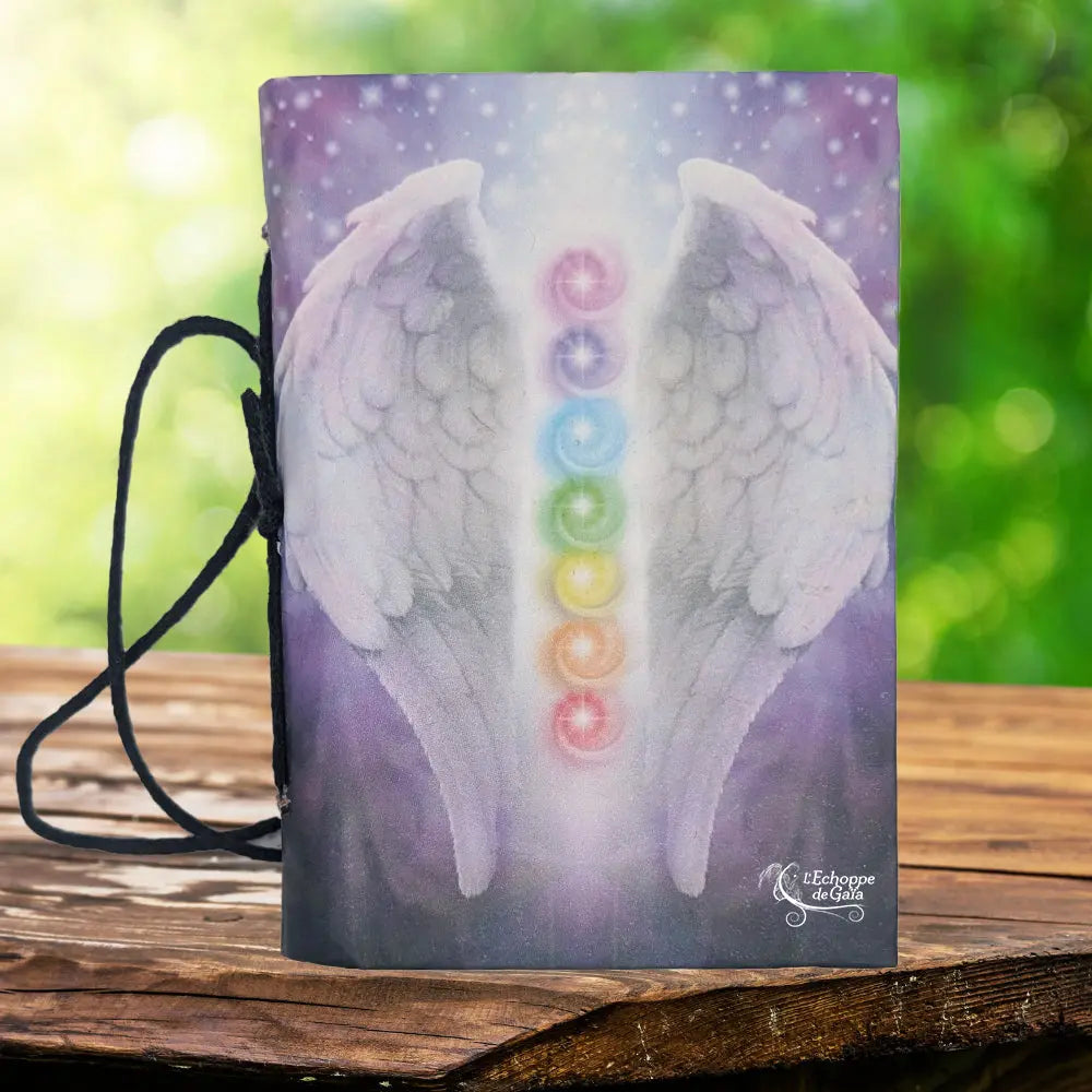 La couverture d'un carnet de notes illustrée d'ailes d'ange blanches et des sept chakras colorés sur un fond violet, muni d'un cordon de fermeture noir et posé sur un support en bois.