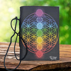 La couverture d'un carnet de notes noir ornée d'une fleur de vie aux couleurs de l'arc-en-ciel et des symboles des sept chakras, muni d'un cordon de fermeture et posé sur un support en bois.