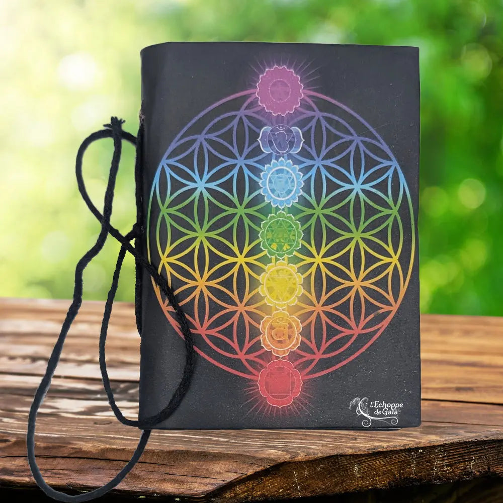 La couverture d'un carnet de notes noir ornée d'une fleur de vie aux couleurs de l'arc-en-ciel et des symboles des sept chakras, muni d'un cordon de fermeture et posé sur un support en bois.