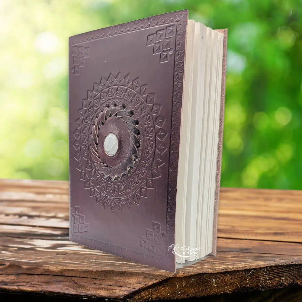La couverture d'un carnet en cuir artisanal marron fermé par deux lanières horizontales, ornée d'une pierre de lune centrale sertie dans une tresse de cuir, sur un support en bois.