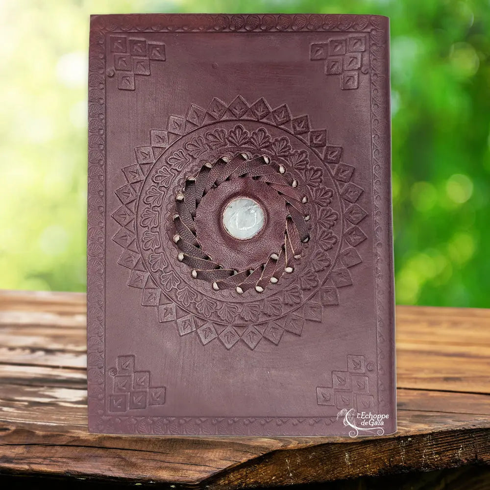 La couverture d'un carnet grimoire en cuir marron ornée de motifs floraux embossés en mandala, centrée sur une pierre de lune sertie dans une tresse de cuir, présentée sur un support en bois.