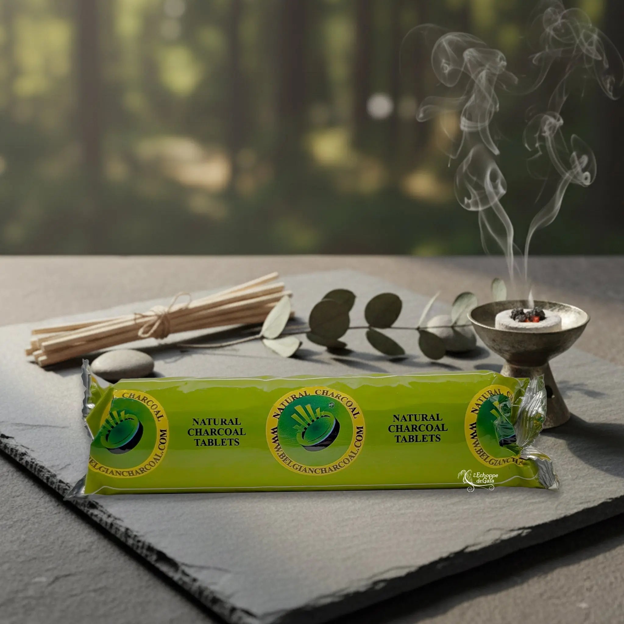 Un rouleau vert de dix pastilles de charbons ardents naturels de 40 mm emballé hermétiquement, posé sur une plaque d'ardoise à côté d'un brûle-encens métallique diffusant de la fumée.