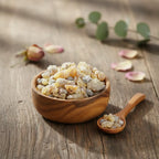 Encens copal en grains clairs dans une coupelle en bois avec petite cuillère sur table en bois, pétales et feuilles en arrière-plan.