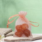Un petit sac en organza pêche contenant des pierres de cornaline, posé sur une pierre plate naturelle devant un arrière-plan vert clair.