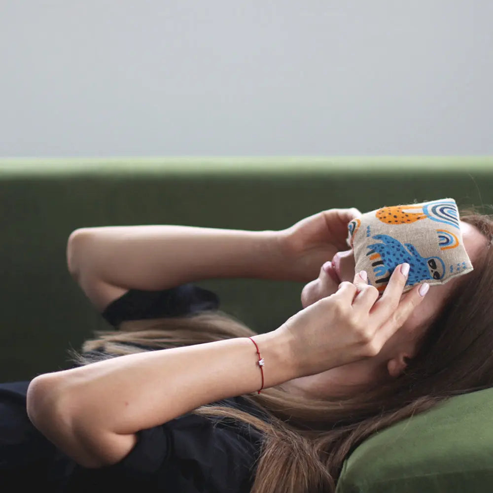 Femme allongée sur un canapé vert, appliquant un coussin chauffant pour les yeux motif paresseux.