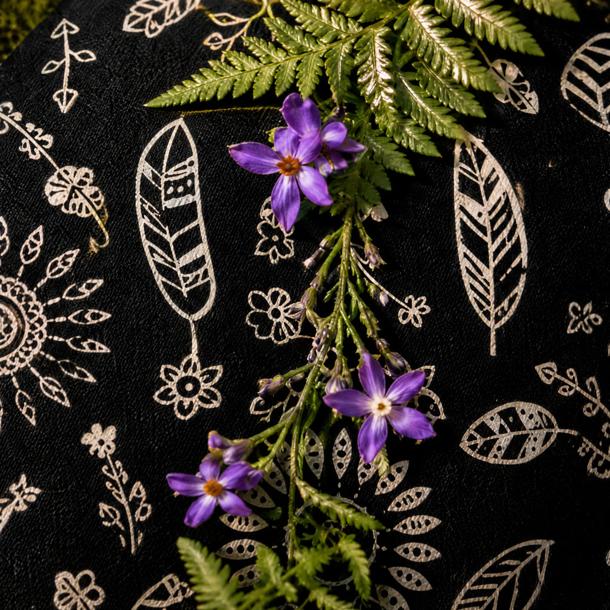 Gros plan sur un coussin chauffant noir aux motifs attrape-rêves et plumes, orné de petites fleurs violettes et de fougères.