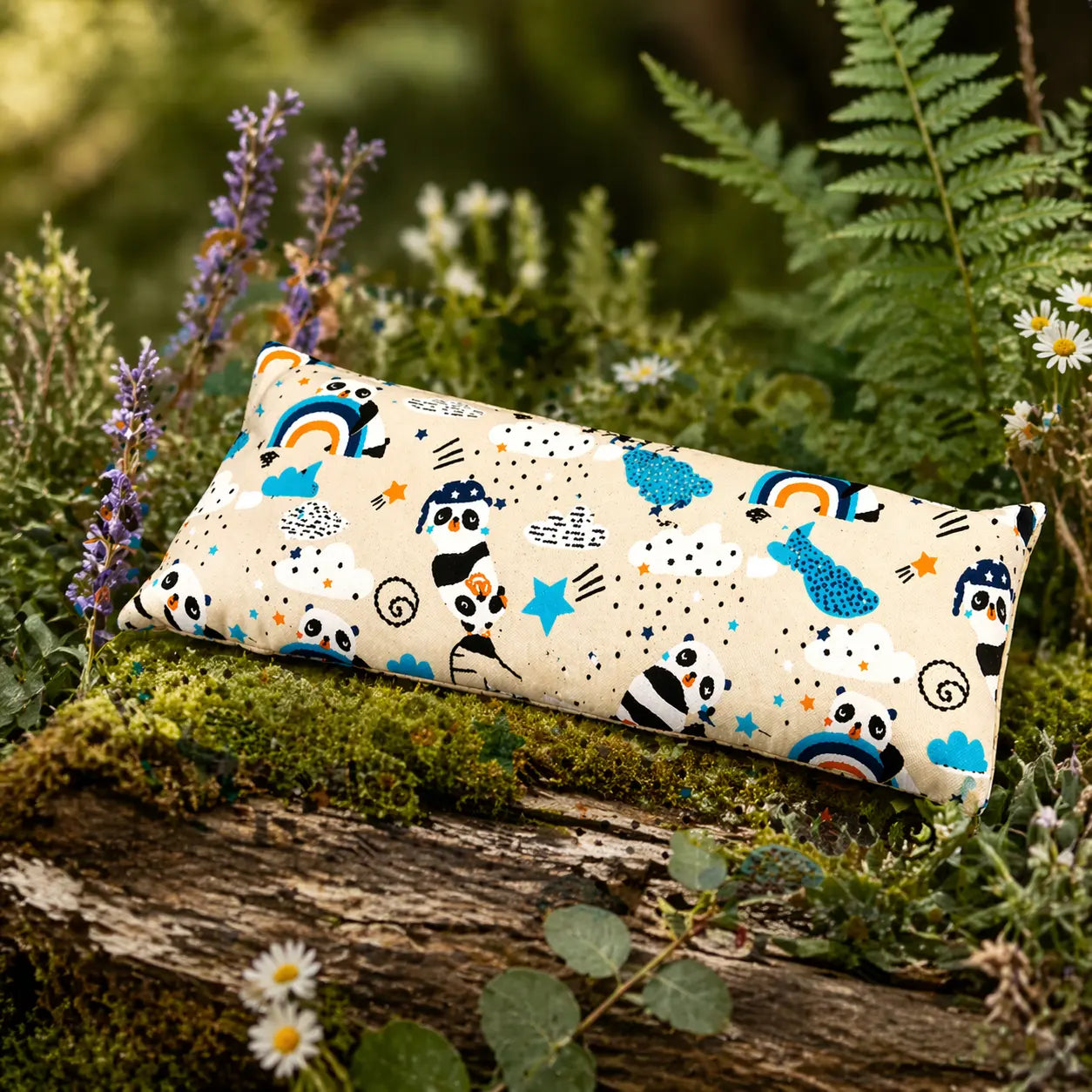 Coussin chauffant rectangulaire aux motifs de pandas, arcs-en-ciel et étoiles, posé sur un tronc d'arbre moussue en pleine nature, entouré de fleurs sauvages et de fougères.